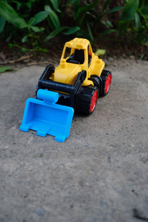 Bright Yellow Toy Construction Vehicle With Blue Scoop Resting on Dirt Near Greenery in Sunny...