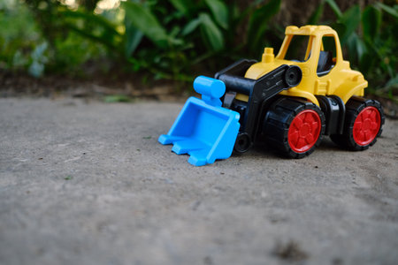 Bright Yellow Toy Construction Vehicle With Blue Scoop Resting on Dirt Near Greenery in Sunny Outdoor Setting.の写真素材