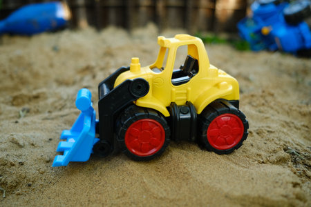 A bright yellow bulldozer toy digs in the sand at a playground filled with colorful plastic toys The...