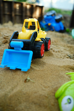 A bright yellow bulldozer toy digs in the sand at a playground filled with colorful plastic toys The...