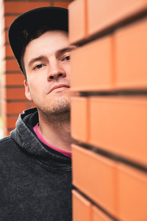 A young man stands partially hidden behind a brick wall peering out with a curious expression in a bright outdoor setting The scene captures a moment of intrigue and urban life.の写真素材