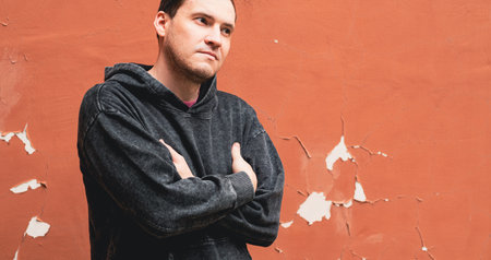 A young man wearing a gray hoodie stands with a serious expression against an orange wall in an urban setting The sunlight highlights his thoughtful demeanor and casual attire.の写真素材