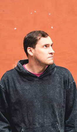 A young man wearing a gray hoodie stands with a serious expression against an orange wall in an urban setting The sunlight highlights his thoughtful demeanor and casual attire.の写真素材