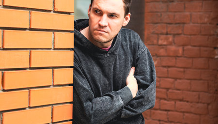 A young man stands with his arms crossed leaning against a warm-toned brick wall He gazes thoughtfully suggesting introspection and contemplation in a vibrant city environment.の写真素材