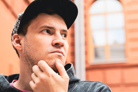 A young man stands in a sunlit courtyard thoughtfully resting his chin on his hand The warm-toned brick walls and tall windows create a vibrant urban atmosphere.の写真素材