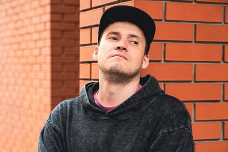 In an urban setting a young man stands confidently against a red brick building He wears a casual hoodie and a cap looking thoughtfully off to the side.の写真素材