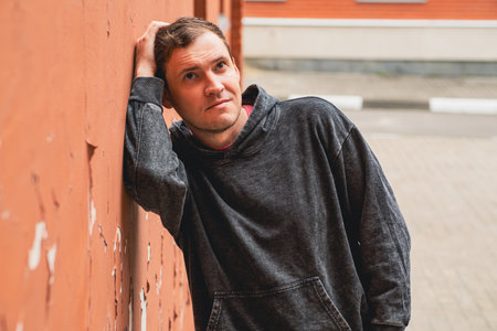 A young man in a dark hoodie leans against a weathered brick wall gazing thoughtfully towards the...