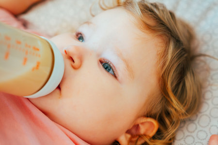 A young child with curly hair is calmly drinking from a bottle while lying on a cozy blanket Sunlight fills the room creating a peaceful atmosphere for the little one.の写真素材