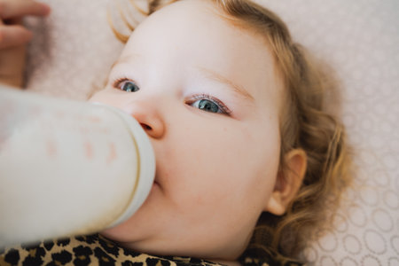 A young child with curly hair is drinking from a bottle propped up comfortably on a soft surface at home showing a relaxed and serene moment during milk time.の写真素材