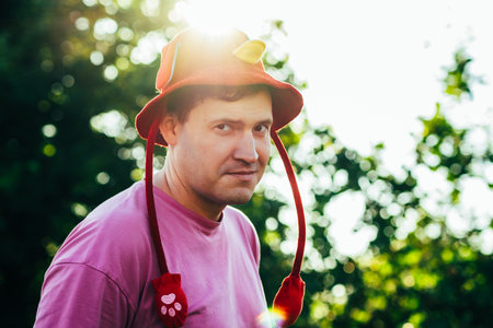Man Wearing a Red and Yellow Playful Hat Enjoys a Sunny Outdoor Moment Surrounded by Trees in a Park.の写真素材