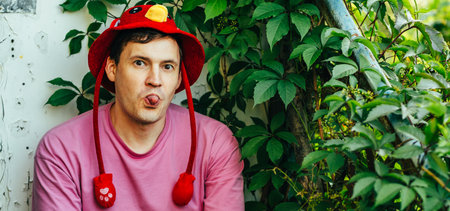 A young man is sitting in a lush garden wearing a colorful hat and a pink shirt while playfully...