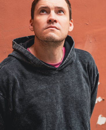 A man gazes upward with a thoughtful expression dressed in a dark hoodie and casual attire He stands...