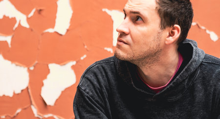 A man wearing a gray hoodie appears lost in thought as he sits against a faded orange wall The wall...