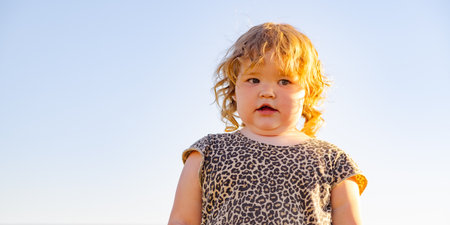 A small child with curly hair stands confidently outside dressed in a leopard print dress The bright sky creates a warm atmosphere capturing a moment of innocenceの写真素材