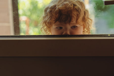 A young child with curly hair looks through a window displaying a sense of curiosity while sunlight brightens the garden outside The outdoor atmosphere is vibrant and livelyの写真素材
