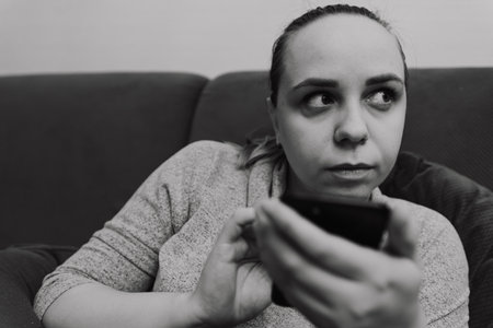 A woman sits on a couch holding a smartphone with a worried expression She appears to be thinking deeply reflecting concern or contemplation in a relaxed home environmentの写真素材