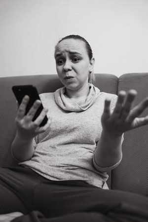 A woman sits on a couch visibly frustrated while looking at her smartphone She gestures with her hands indicating talk or disbelief The atmosphere is casual and intimateの写真素材