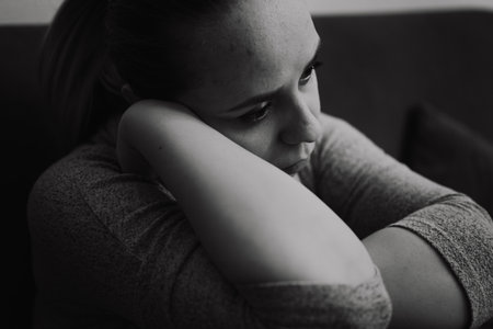 The image captures a woman resting her chin on her arm while sitting on a couch surrounded by a subdued atmosphere that enhances her thoughtful demeanor and pensive moodの写真素材