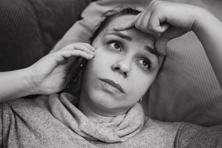 A woman is lying on a couch with her hand on her forehead appearing deep in thought as she talks on the phone The indoor setting is inviting and comfortableの写真素材