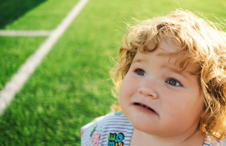 A young child with curly hair gazes up curiously while standing on a vibrant green field Sunlight...