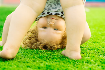 A young girl with curly hair enjoys a playful moment outside doing a handstand on bright green grass...