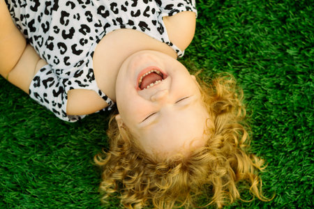 A cheerful young girl lies on soft green grass laughing joyfully as she delights in the playful moments of her day while enjoying warm sunshine outdoorsの写真素材
