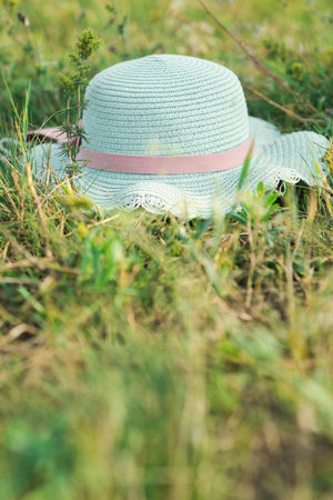 A stylish straw hat with a pink ribbon gently lies on soft green grass The surrounding field is vibrant with wildflowers and tall grasses under a clear blue skyの写真素材