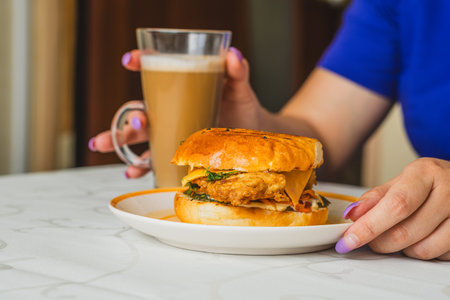 A person enjoys a crispy chicken sandwich placed on a white plate alongside a cup of warm coffee The soft bun and fresh ingredients highlight a satisfying breakfast experienceの写真素材