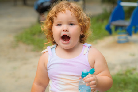 A joyful young girl with curly hair is seen playing in a park. She holds a bottle of water while expressing excitement, enjoying her time outdoors during a warm day.の写真素材