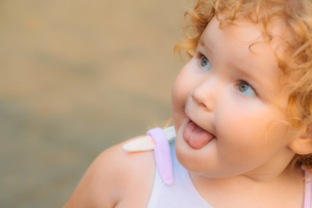 A young child with curly hair playfully sticks out her tongue while laughing. Bright sunlight...