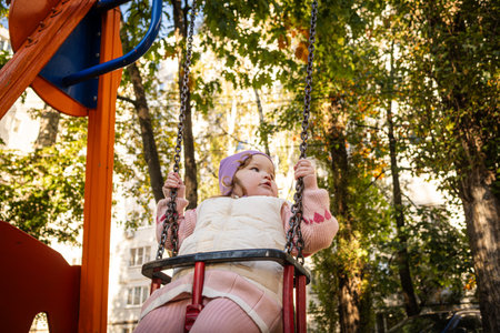 A joyful toddler swings in the park, wearing a purple hat and a cozy vest. Bright autumn leaves create a warm atmosphere, while laughter fills the air.の写真素材