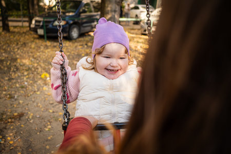 A joyful toddler swings in the park, wearing a purple hat and a cozy vest. Bright autumn leaves create a warm atmosphere, while laughter fills the air.の写真素材