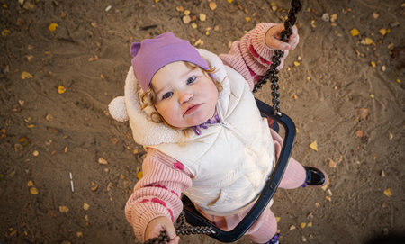 A joyful toddler swings in the park, wearing a purple hat and a cozy vest. Bright autumn leaves create a warm atmosphere, while laughter fills the air.の写真素材