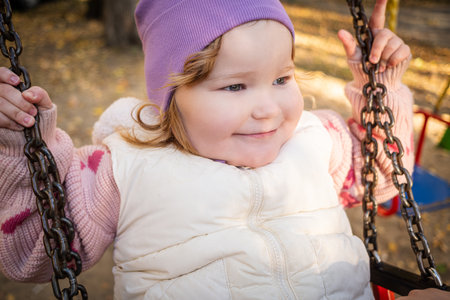 A joyful toddler swings in the park, wearing a purple hat and a cozy vest. Bright autumn leaves create a warm atmosphere, while laughter fills the air.の写真素材