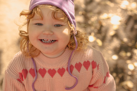 A cheerful toddler with curly hair and a purple hat smiles brightly outside, surrounded by a warm, sunny atmosphere and golden leaves in the background.の写真素材