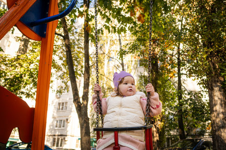 A joyful toddler swings in the park, wearing a purple hat and a cozy vest. Bright autumn leaves create a warm atmosphere, while laughter fills the air.の写真素材