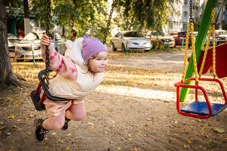 A joyful toddler swings in the park, wearing a purple hat and a cozy vest. Bright autumn leaves create a warm atmosphere, while laughter fills the air.の写真素材