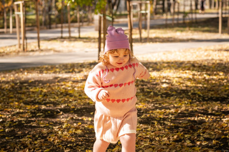 A young child with curly hair runs joyfully through a park covered in colorful autumn leaves. She wears a cozy sweater and a cute hat, enjoying the beautiful sunny day.の写真素材