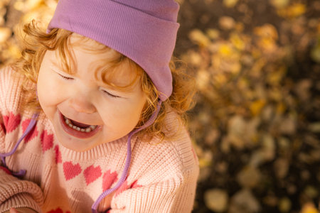 Child in purple hat surrounded by autumn leaves in a park on a sunny dayの写真素材