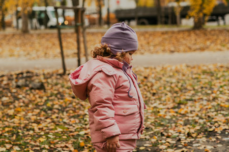 A small child enjoys the cool autumn day in a park. She wears a pink coat and purple hat, exploring the ground covered in vibrant yellow and orange leaves.の写真素材