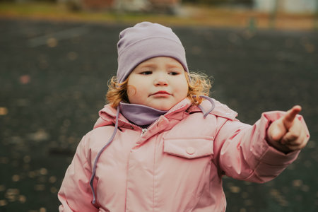 A young child wearing a pink coat and purple hat points at something interesting while playing in a park. The leaves are scattered around, showing autumn's arrival.の写真素材
