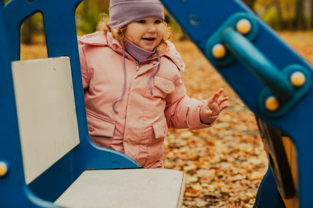 A young girl in a pink jacket and purple hat plays joyfully at a playground. The vibrant colors of autumn leaves create a beautiful backdrop for her fun activities.の写真素材