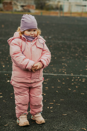 A small child stands alone on a playground with a sad expression, dressed warmly in a pink coat and hat. The playground is empty, and the cold wind is evident, making her feel upset.の写真素材