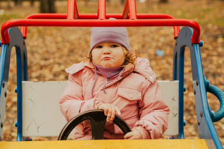 A little girl with curly hair, dressed in a pink coat and purple hat, sits in a toy car at a park surrounded by fallen leaves. It sa chilly autumn day with a playful atmosphere.の写真素材