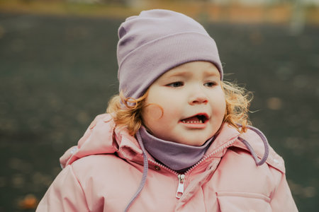 A young girl with curly hair wearing a purple hat and a pink coat smiles while playing outdoors in a park on a chilly autumn day filled with fallen leaves.の写真素材