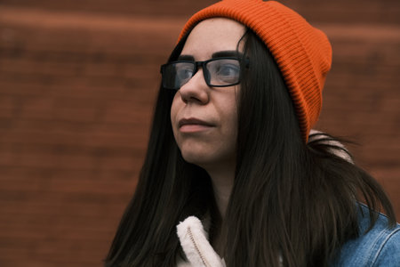 A young woman stands against a textured brick wall wearing a cozy denim jacket and a vibrant orange beanie She gazes upward showcasing a stylish look that reflects urban fashionの写真素材