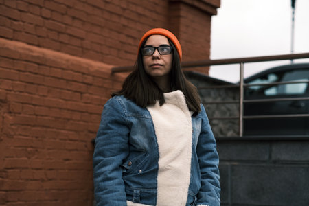 Fashionable Young Woman Poses With Confidence in a Denim Jacket and Orange Beanie Against a Brick...