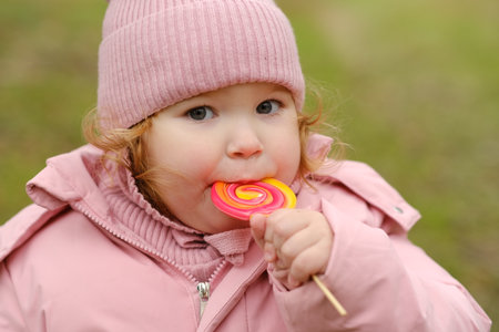 Child Enjoys Colorful Lollipop While Wearing Warm Clothing on a Cool Overcast Day Outdoors in a Park Settingの写真素材