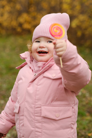 A cheerful kid stands outside in a bright pink coat and matching hat happily holding a swirl lollipop against a backdrop of colorful autumn leaves The scene captures a moment of joyの写真素材