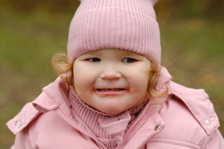 A young child dressed in a pink coat and hat is outside in a green area The child seems to show surprise or concern while enjoying the day The weather appears cool and the environment is naturalの写真素材